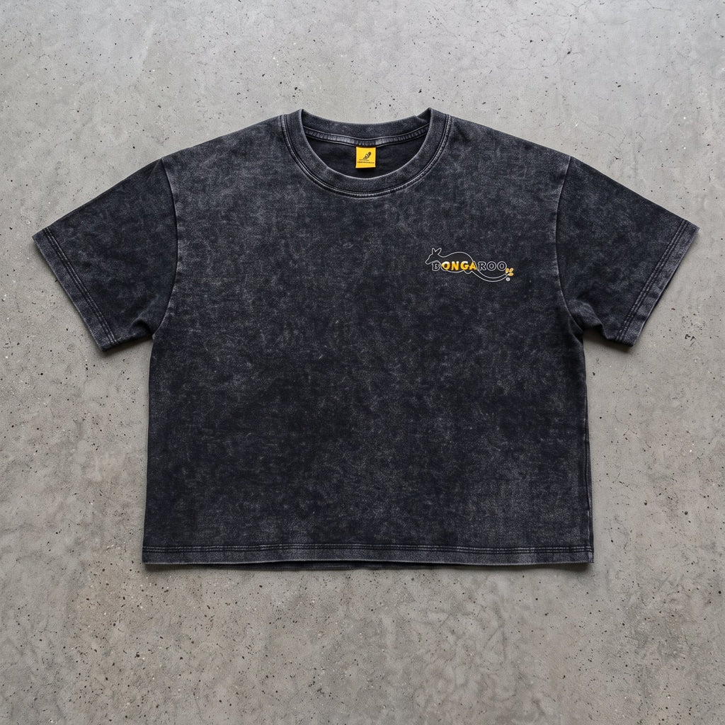 A dark gray acid-wash cropped t-shirt with a small yellow and black graphic on the left chest, laid flat on a textured gray concrete surface.