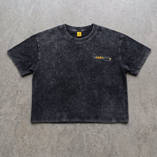 A dark gray acid-wash cropped t-shirt with a small yellow and black graphic on the left chest, laid flat on a textured gray concrete surface.